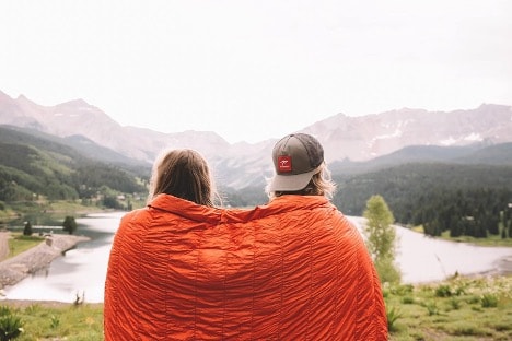 Two people sitting on a hill, in a blanket looking at a lake