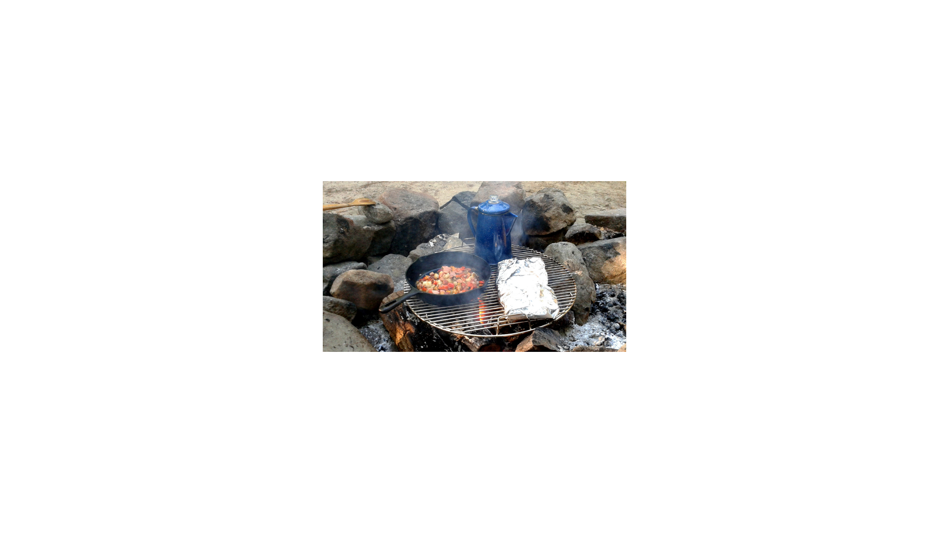 Food being cooked over a campfire, foil packed food and a cast iron skillet, and a kettle.